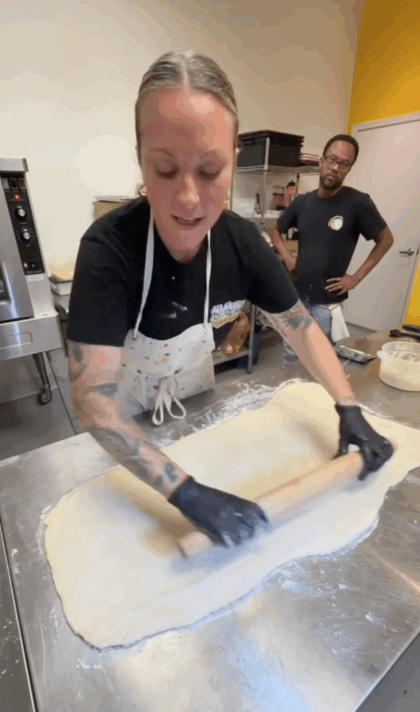 Lindsay baking rolling cinnamon rolls while Lawrence oversees from behind.